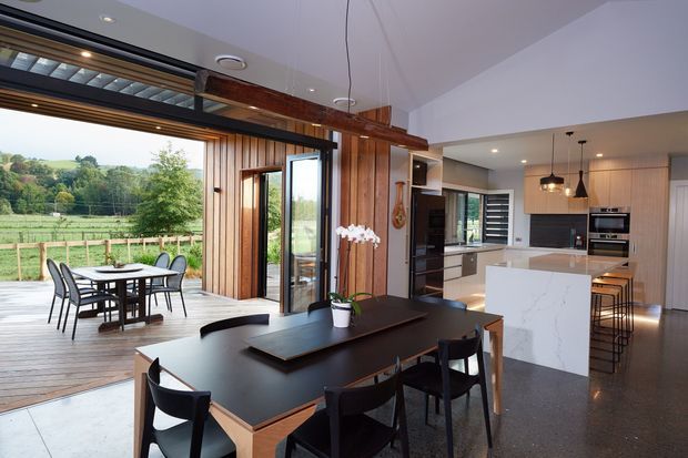 Open-plan dining and kitchen interior with a large dark dining table, black chairs, and a white island bench. Full-height sliding glass doors open onto a timber deck overlooking a green rural landscape. The room features warm vertical timber wall cladding, polished concrete flooring, contemporary cabinetry, pendant lights, and a timber-framed opening with exposed structural detailing.