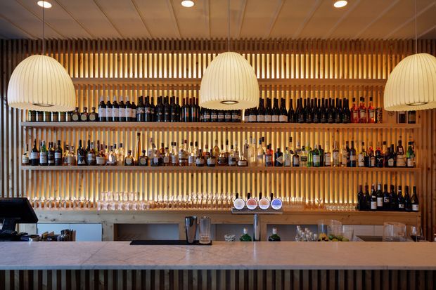 A warmly lit contemporary bar interior with a long pale stone or composite counter in the foreground, extensive back-bar shelving filled with bottles and glassware, and a feature wall of closely spaced vertical timber slats. Three large ribbed pendant lights hang above the bar, complemented by recessed ceiling lighting. The space has a refined hospitality fit-out with strong timber detailing and warm ambient illumination.