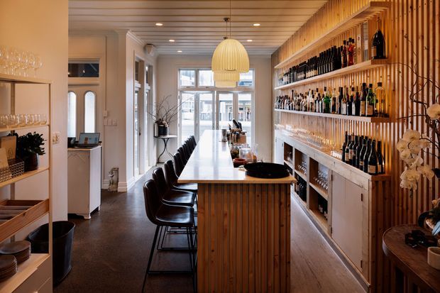 A narrow, warmly lit commercial bar or hospitality space with a long central timber-clad counter and bar seating. The right wall features extensive open bottle shelving, glassware, cabinetry and vertical timber battens, while a large pendant light hangs above the counter. Dark flooring, white walls, and tall glazing create a refined, intimate atmosphere.