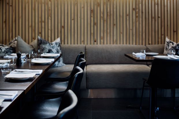 A refined restaurant dining area with dark wood tables set with white crockery and glassware, black upholstered dining chairs, and a long grey upholstered banquette. The backdrop features a full-height vertical timber slat wall, creating a warm yet moody contemporary hospitality interior.