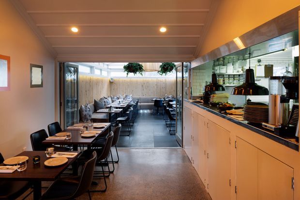 A warm, narrow contemporary restaurant interior viewed from the dining area toward a glazed outdoor courtyard. Dark dining tables and chairs line the left side, while a long service counter with pale cabinetry, black benchtop, pendant heat lamps, shelving, and a tiled backsplash occupies the right. The space features a coffered or panelled ceiling with recessed lighting, polished concrete flooring, and extensive glazing opening to an outdoor dining area.