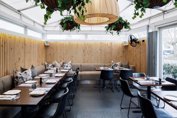 Bright contemporary restaurant dining area with rows of dark timber tables and black chairs, upholstered banquette seating along a vertical timber slat wall, large windows, and abundant hanging greenery. A prominent woven pendant light and suspended planters add warmth and visual interest to the otherwise neutral fit-out.