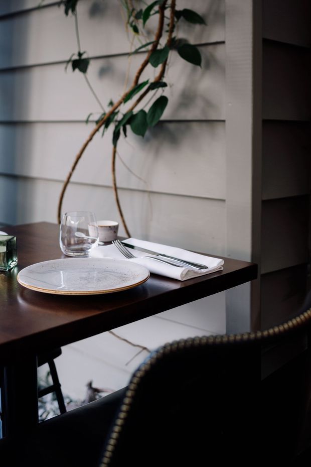A moody close-up of a dining setting in a compact interior. A dark timber table is set with a ceramic plate, folded napkin, cutlery, drinking glass and small cup, with a dark upholstered chair in the foreground. White horizontal wall panelling and a tall climbing indoor plant form the backdrop, while open shelving is visible at right.