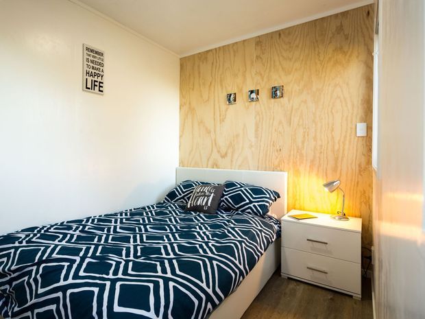 Compact bedroom with a double bed dressed in navy-and-white geometric bedding, a pale upholstered headboard, a warm plywood feature wall, white painted walls, a small white bedside unit, and a desk lamp. The room has a simple, casual contemporary aesthetic with warm timber flooring and minimal wall decor.