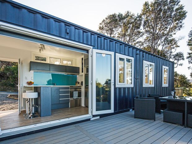 Exterior view of a compact dark blue corrugated-clad dwelling or studio opening onto a broad timber-look deck. The open glazed wall reveals a small contemporary kitchen with blue cabinetry, a turquoise splashback, stainless-steel appliances and bar seating, while outdoor dining furniture occupies the deck. Tall trees are visible behind the building.