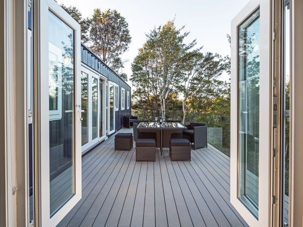 A spacious outdoor timber deck viewed through open glass doors, furnished with a dark woven dining table and chairs. The deck is enclosed by glass balustrading and bordered by a dark-clad building with extensive white-framed glazing, surrounded by mature trees.