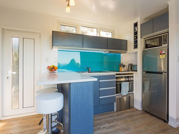 A compact contemporary kitchen with blue-grey wood-grain cabinetry, a glossy turquoise glass splashback, white benchtops, stainless-steel appliances, and timber-look flooring. The kitchen includes a small peninsula with a white bar stool, integrated microwave, oven, cooktop, refrigerator, sink, and under-cabinet lighting. White walls, a glazed back door, and upper windows make the space bright and functional.