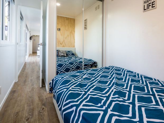 A compact bedroom viewed toward a narrow hallway, with a single bed covered in bold blue-and-white geometric bedding. Mirrored wardrobe doors reflect the bed and room, while a plywood feature wall adds warmth. White walls, doors, trim, and cabinetry create a bright, simple interior, with timber-look flooring extending through the room and hall.
