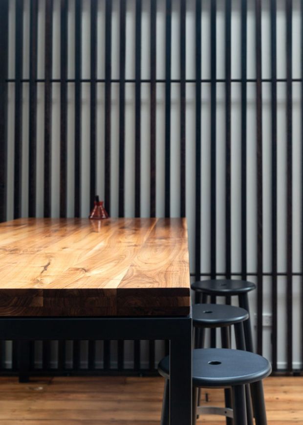 A close-up interior view of a dark dining table with a thick warm timber top and black base, paired with black stools. The backdrop is a dramatic vertical black-and-light slatted wall, with timber flooring and a small decorative object on the table. The composition has a modern, high-contrast, refined character.