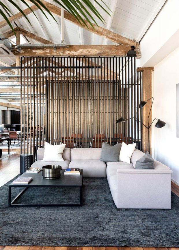 A refined open-plan living room with a large pale sectional sofa, dark rectangular coffee table, and charcoal area rug. The space features exposed weathered timber beams, a high white pitched ceiling, timber flooring, and a dramatic black vertical-slat screen separating the lounge from the dining or work area. A sculptural multi-arm floor lamp and indoor palm add warmth to the industrial, loft-like setting.