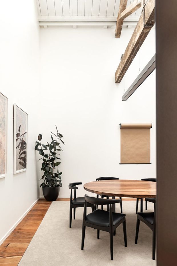 Bright dining room with a round mid-tone timber dining table, black dining chairs, a large neutral rug, framed botanical artwork, and a potted rubber plant. The room has white walls and a white-lined high ceiling with exposed rustic timber beams and a suspended dark structural beam, creating a refined warehouse or loft-like character.