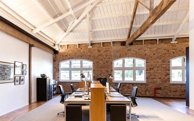 Bright open-plan office set within a converted brick industrial-style space, featuring exposed brick walls, white-painted vaulted ceiling with exposed timber and painted structural beams, large arched-top windows, timber flooring, and a central cluster of desks and office chairs.