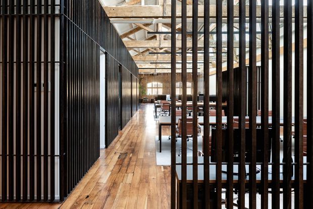 An elongated industrial-style commercial interior with a timber floor, exposed timber roof structure, and rows of dark vertical metal screens dividing a central circulation path from furnished work or dining areas. Natural light enters through large windows at the far end, creating a warm contrast between the wood surfaces and black metal partitions.