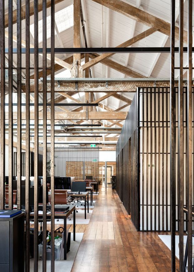 A long, open-plan commercial interior with exposed timber roof trusses and beams, white pitched ceiling panels, warm timber flooring, and extensive black metal vertical screens and partitions. Workstations and lounge-style office furniture line the corridor, creating an industrial warehouse-conversion character with strong linear perspective and abundant natural light.