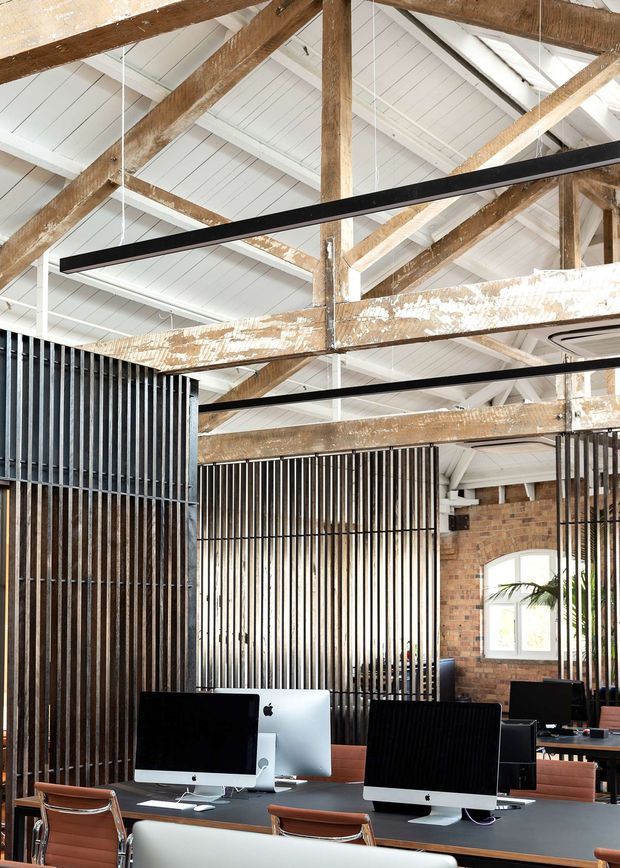 A bright, open-plan office interior set within a reused industrial-style building. Exposed timber roof trusses and beams sit beneath a white pitched ceiling, while black linear pendant lights hang above shared workstations. Vertical black metal screening partitions define the workspace, with exposed brick walls and large windows adding texture and natural light. Multiple computer workstations and office desks occupy the foreground.