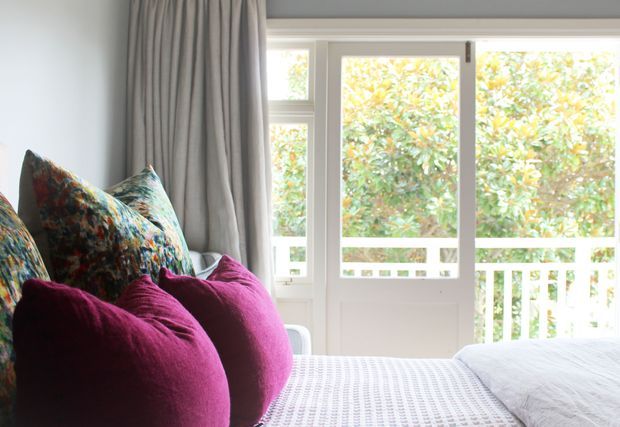 Bright, cozy bedroom corner with a bed in the foreground, layered textured magenta and patterned pillows, soft grey floor-length curtains, and large white-framed glazed doors or windows opening toward a leafy exterior and balcony railing. The room has a relaxed, colorful, contemporary feel with abundant natural light.