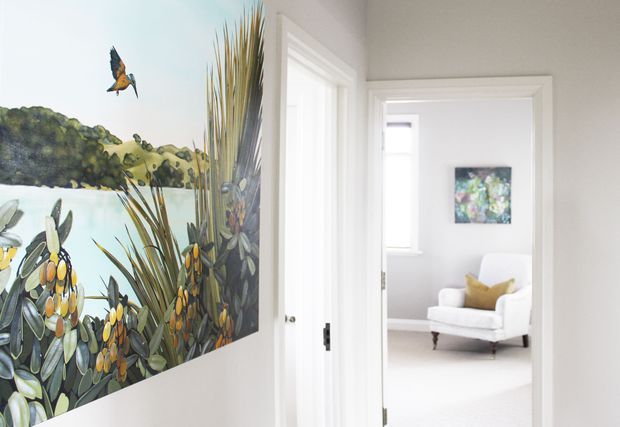 Bright, refined interior view of a white-painted hallway or transition space opening into a light-filled living room. The foreground wall features a large colourful nature mural with foliage and a bird, while the adjoining room contains a white upholstered armchair, a small mustard cushion, artwork, and pale finishes. White trim, door frames, and abundant natural light create a clean, airy atmosphere.