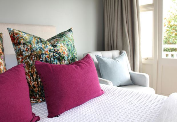 A close-up of a bright, cozy bedroom seating or bed area with a pale upholstered headboard, layered patterned and magenta cushions, a pale blue armchair, a white textured bedspread, and full-height grey curtains beside a glazed balcony door. The styling is colourful and refined with abundant natural light.