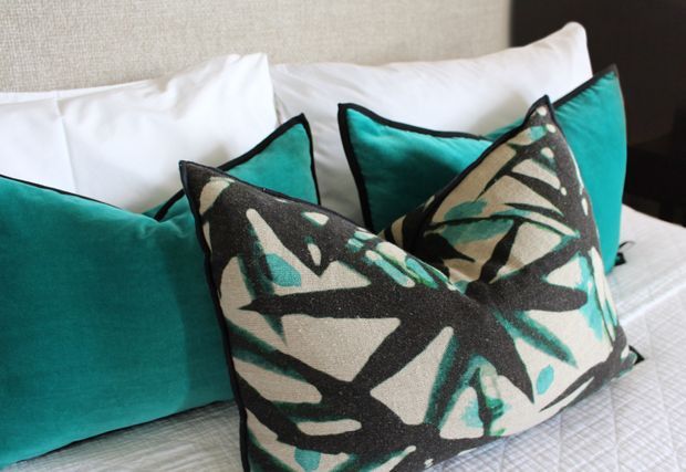 Close-up of a neatly styled bed with layered white pillows and vivid turquoise accent cushions, including a prominent black, cream, and turquoise abstract botanical-patterned cushion. A neutral upholstered headboard and light bedding create a refined, high-contrast bedroom setting.