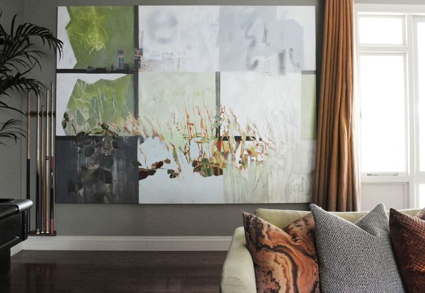 A refined living room corner with a large abstract multi-panel artwork on a dark grey wall, a pale upholstered sofa with patterned cushions, dark timber flooring, tall indoor plants, and full-height warm drapery beside a bright window. The room has a contemporary, art-led and subdued atmosphere.