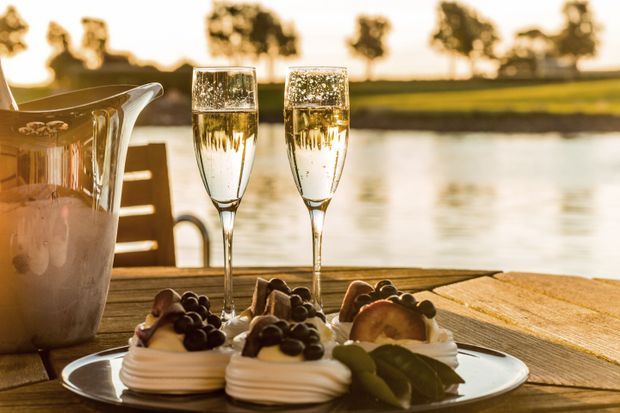 A sunlit outdoor dining setting beside calm water, with a wooden table holding two champagne flutes, a metal ice bucket, and plated desserts topped with fruit. The scene has warm golden-hour lighting and a relaxed, refined entertaining atmosphere.