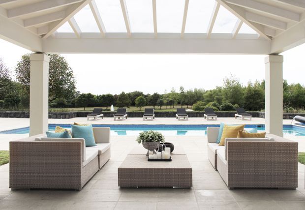 A bright covered outdoor lounge with a white timber-framed roof and translucent panels, furnished with two facing wicker sofas, pale cushions, and a central wicker coffee table. It opens directly onto a paved pool terrace with a long swimming pool and landscaped garden beyond.