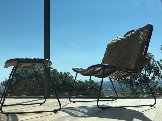 A sunlit outdoor terrace or balcony with two mid-century-style sling lounge chairs, one draped with a knitted throw. The chairs sit on pale decking or paving beside full-height glazing, with leafy vegetation and a distant landscape visible beyond. Strong sunlight and long shadows create a relaxed scenic setting.