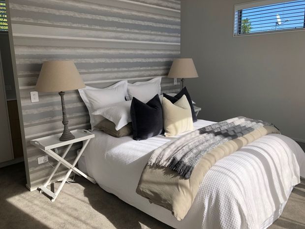 A styled bedroom with a neatly made bed, layered white and neutral bedding, dark accent cushions, bedside tables, and matching table lamps. The room features a horizontally textured grey feature wall, neutral painted walls, carpet flooring, and a high horizontal louvre window.