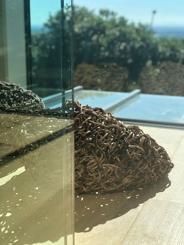 A sunlit outdoor terrace or poolside area viewed beside a clear glass balustrade. A large woven natural-fibre cushion or sculptural outdoor furnishing sits on pale timber decking, with a blue swimming pool and blurred greenery beyond. Strong reflections and shadows create a warm, tranquil atmosphere.