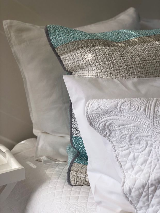 Close-up of a neatly made bed with layered white pillows and quilted bedding, featuring a textured teal and taupe decorative cushion and a white embroidered cushion. The scene has soft natural light and a calm, refined bedroom styling.