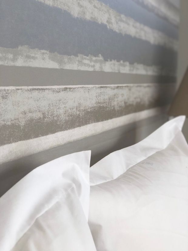 Close-up interior view of a bed with crisp white bedding in the foreground and a textured, horizontally striped grey feature wall behind it. The image has a soft, neutral, contemporary bedroom aesthetic with minimal visible furnishings.