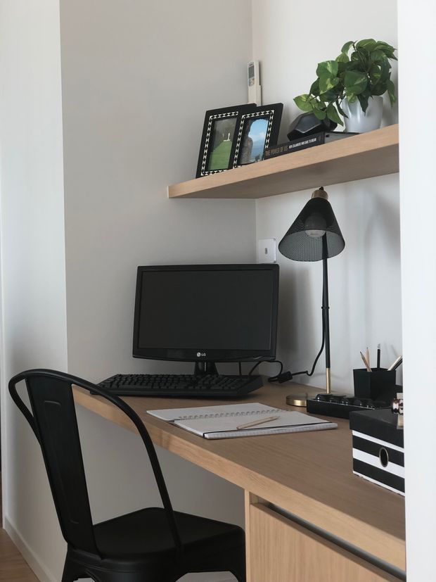 Compact home office nook with a built-in light-wood desk, black computer monitor and keyboard, black metal chair, desk lamp, stationery, and a floating timber shelf displaying framed photos and a potted plant. The space has white walls and a clean, functional contemporary feel.