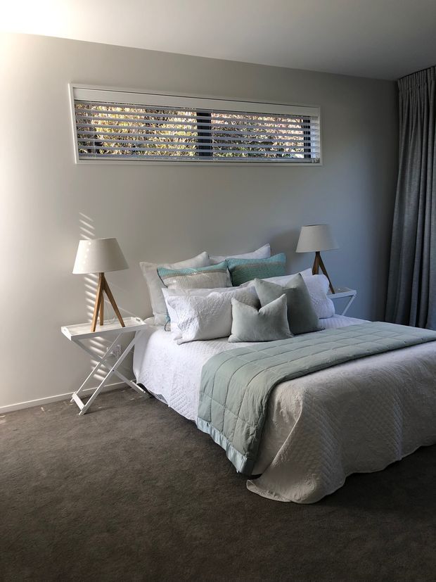 A softly styled bedroom with a neatly made bed layered in white bedding and pale blue-grey cushions and quilt. Matching tripod lamps sit on small white folding side tables, while a horizontal louvred window and full-height dark curtains frame the room. The space has neutral painted walls and dark carpet, with gentle natural light creating a calm atmosphere.