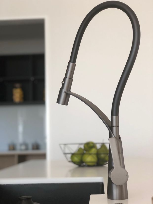 Close-up product-style photograph of a tall gooseneck kitchen mixer tap with a pull-out spray head and side control lever, finished in dark brushed metal. It is installed at a kitchen island or benchtop, with a black sink edge visible in the foreground and blurred kitchen shelving and a fruit bowl in the background.