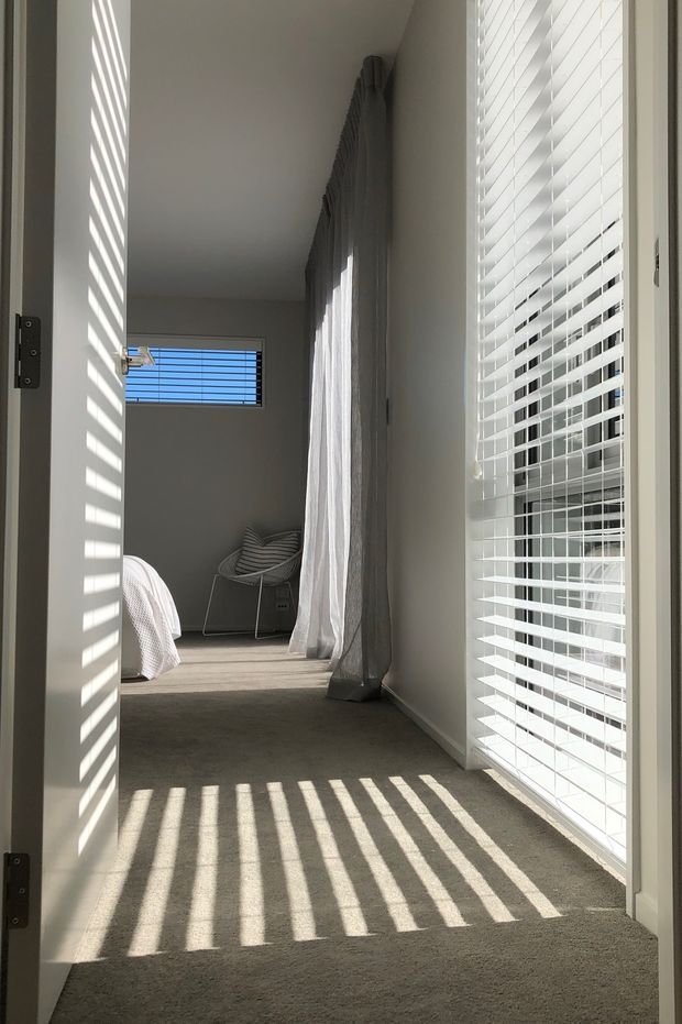 A bright, narrow bedroom viewed through an open doorway, with soft grey carpet, white walls, sheer grey curtains, and large windows fitted with white horizontal blinds. Strong sunlight casts striped shadows across the carpet and walls. A partial bed and a small upholstered chair are visible at the far end.