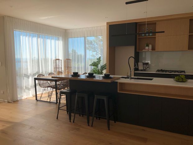 Open-plan contemporary kitchen and dining area with a dark island incorporating three stools, pale timber flooring, light timber cabinetry, dark lower cabinetry, and a slim stone-look benchtop. A dining table sits beside large sheer-curtained windows, with simple pendant lighting and integrated shelving.