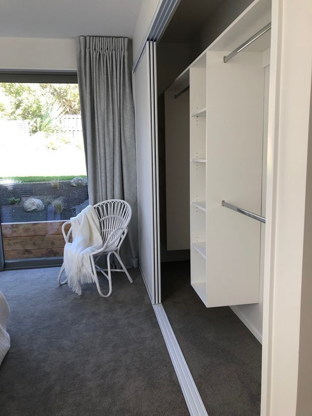 A bright built-in walk-in wardrobe beside a carpeted bedroom or dressing area. White cabinetry with open shelving and chrome hanging rails is concealed behind sliding doors. A large window with grey curtains brings in natural light, and a white woven lounge chair with a throw sits nearby.