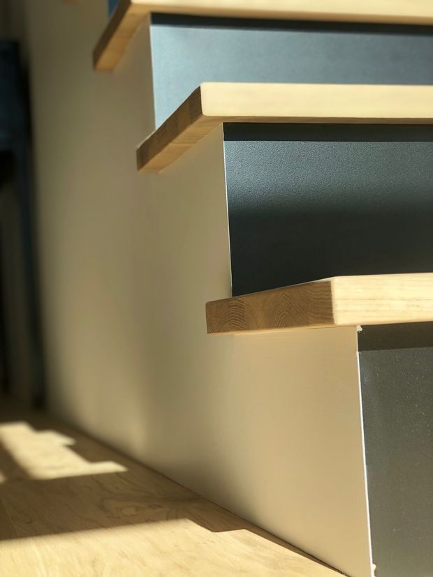 Close-up architectural detail of floating timber shelves or stair treads projecting from dark charcoal and light grey wall panels. Warm natural wood grain, crisp geometric edges, and strong sunlight create pronounced shadows across the pale timber floor and wall surfaces.