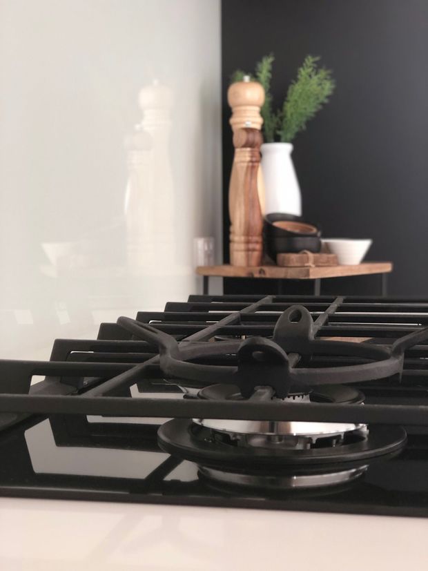 Close-up view of a modern black gas cooktop with cast-iron grates set into a light benchtop. A dark feature wall, small timber shelf, wooden pepper mill, white vase, greenery and kitchen accessories appear in the background.