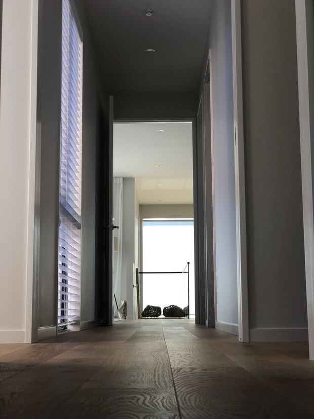 A low-angle view down a narrow, modern interior hallway with dark timber flooring, pale walls, white trim, a tall louvred window on the left, and open doorways leading toward a bright glazed area at the far end. The corridor has a restrained, contemporary feel with recessed ceiling lights and strong contrast between the darker foreground and daylight beyond.