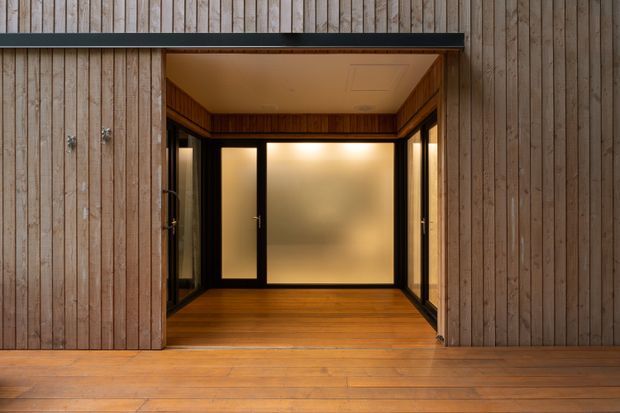 A warm, compact timber-lined entry or enclosed porch opening from a timber-clad exterior deck. Vertical weathered timber boards frame black-framed frosted glazing on three sides, with a timber floor and timber-lined ceiling creating a sheltered, minimalist threshold space.