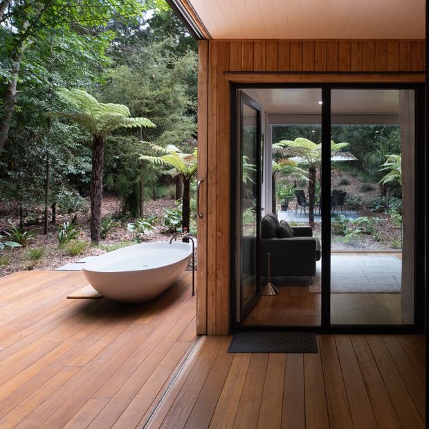 A warm timber-clad indoor-outdoor bathing area opening onto a forested deck. A freestanding white oval bathtub sits on the timber decking beside a black-framed glazed sliding door, with a compact living area visible through the glass. Dense native-looking vegetation and tree ferns surround the private outdoor setting.