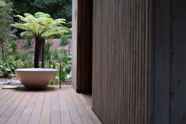A calm, wood-lined bathroom opening onto a lush fern garden. A freestanding sculptural bathtub sits on a timber deck or floor beside a slim dark bath tap, with vertical ribbed timber wall cladding and a deep open doorway framing the greenery.