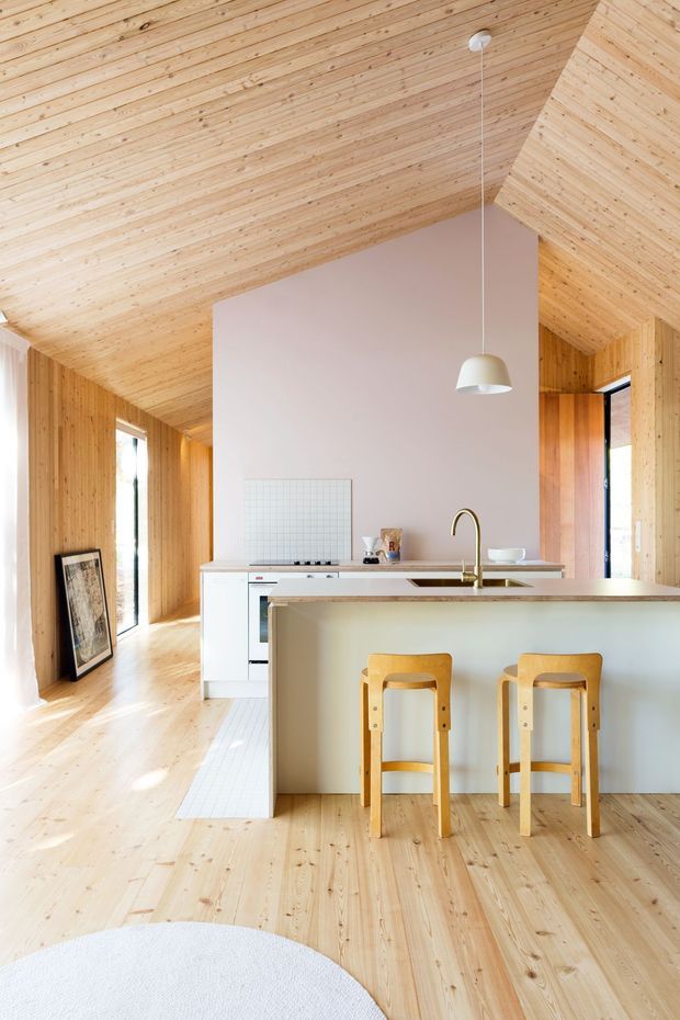 Bright open-plan kitchen with a large pale island and integrated sink, white cabinetry, brass tapware, and two timber bar stools. The room is enveloped in warm light timber flooring, wall panelling, and a pitched timber-lined ceiling, with a muted blush feature wall and minimalist pendant light.
