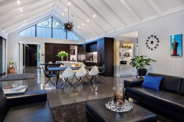Bright open-plan contemporary living and dining area with a kitchen, polished concrete flooring, black leather seating, and a dramatic white vaulted ceiling with exposed beams. Large triangular clerestory glazing fills the room with natural light, while dark timber cabinetry and a sculptural pendant add contrast.