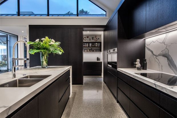 Contemporary kitchen with a large island, integrated sink and brushed metal tap, dark flat-panel cabinetry, polished stone benchtops, and a dramatic veined stone splashback. High clerestory glazing brings in natural light, while a walk-in pantry or beverage station is visible at the rear. The polished terrazzo-like floor and black cabinetry create a refined, high-contrast interior.