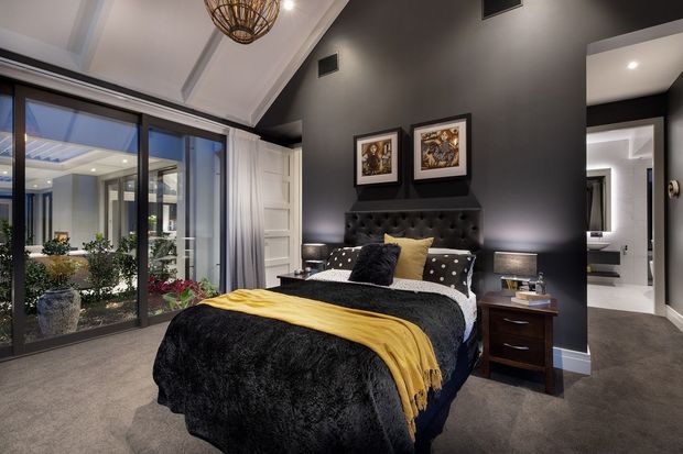 A sophisticated contemporary bedroom with a black upholstered tufted bedhead, dark charcoal walls and ceiling accents, layered black-and-white bedding, and a mustard-yellow throw and cushions. The room features bedside tables, framed artwork, carpeted flooring, recessed lighting, and large sliding glass doors opening to a planted courtyard or garden. A hallway or adjoining bathroom area is visible through the opening on the right.