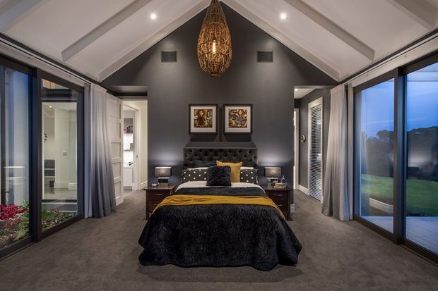 A sophisticated master bedroom with a vaulted white-beamed ceiling, dark charcoal feature wall, and expansive floor-to-ceiling sliding glass doors on both sides. The room features a centred bed with a tufted dark headboard, layered black, grey, and mustard bedding, matching bedside tables and lamps, framed artwork, plush grey carpet, sheer curtains, and a large woven pendant light. Outdoor greenery and views are visible through the glazing.