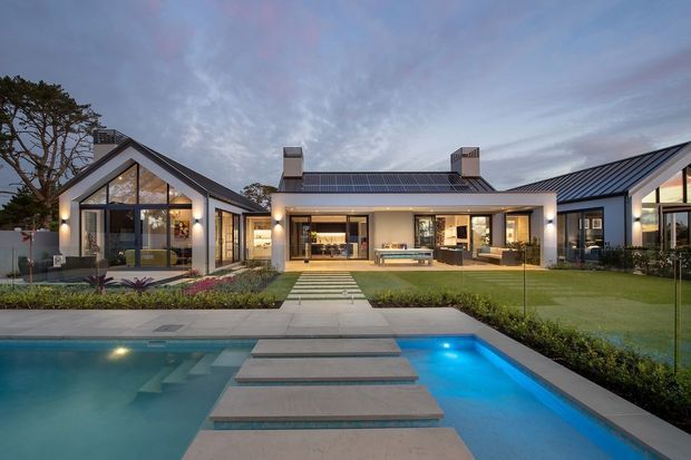 A contemporary single-storey residence arranged around a landscaped courtyard and swimming pool. The home features multiple gabled volumes, dark metal pitched roofs, extensive floor-to-ceiling glazing, pale rendered walls, and a covered outdoor living area opening toward the pool. Stepping stones cross the lawn and pool, with integrated landscape planting and evening architectural lighting.