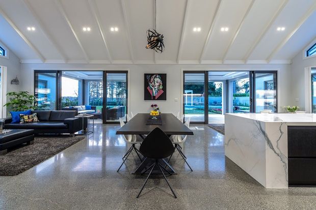 Open-plan contemporary kitchen, dining and living area with a high white pitched-panel ceiling, polished concrete or terrazzo flooring, expansive black-framed glazing, and views toward a swimming pool and landscaped garden. A marble-look island with dark cabinetry anchors the kitchen, while a dark dining table and black lounge furniture create contrast.